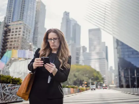 A woman checking her phone on a sunny New York street lined with tall towers