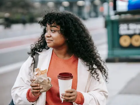 A woman with curly hair holding a coffee beside a subway entrance on a city street