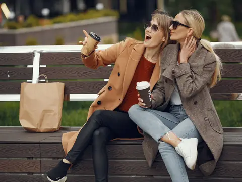 Two friends in warm coats laughing over coffee on a park bench in autumn